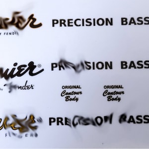 May include: A white sheet with multiple instances of the "Squier" and "Fender" logos, along with "Precision Bass" text. The logos are in black and gold, and the text "Original Contour Body" is also present.