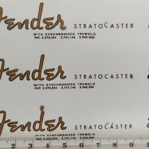 Puede incluir: Tres calcomanías Fender Stratocaster con escritura dorada y texto negro. Cada calcomanía incluye el logotipo de Fender, el nombre del modelo Stratocaster e información de patente. También está presente la frase "Original Contour Body".