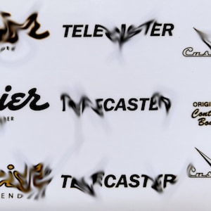 May include: A close-up of three guitar headstock decals. The top decal is gold and says "Squier by Fender" and "Telecaster". The middle decal is black and says "Squier by Fender" and "Telecaster". The bottom decal is gold and says "Squier by Fender" and "Telecaster".