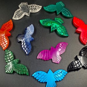 May include: A collection of ten bird-shaped carvings arranged in a circular pattern. The carvings are made in various colors including clear, dark green, bright green, orange, blue, red, magenta, black, and turquoise, each with etched wing details.