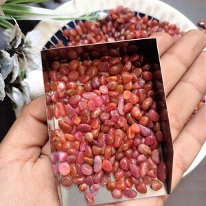 May include: A collection of small, polished, reddish-pink gemstones. The stones are held in a small, square, metal container, with more stones visible in the background. The gemstones vary in color from light pink to deep red.