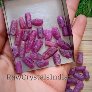 May include: A collection of raw ruby crystals in various shades of pink and purple. The crystals are arranged in a metal tray and are approximately 1-2 cm in length.