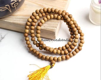 May include: A wooden mala necklace with a yellow tassel. The necklace is made of small, round, light brown beads. The necklace is lying on a wooden surface. A small jar and a singing bowl are in the background.