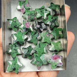 May include: A collection of star-shaped gemstones made of green zoisite with inclusions of pink ruby and black hornblende. The stones are faceted and arranged in a metal tray, displaying a mix of vibrant green, deep pink, and dark black colors.
