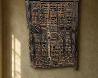 May include: A round dark wood dining table with a central pedestal base, two wooden chairs with woven seats, and a large rectangular carved wooden wall panel featuring geometric patterns and human figures mounted above the table.