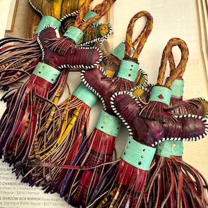 May include: A close-up of several colorful leather tassels with a braided leather loop at the top. The tassels are made of different colors of leather, including red, yellow, green, and blue. The tassels are arranged in a pile, with some of them overlapping. The tassels are a type of decorative trim that can be used on clothing, bags, or other items.