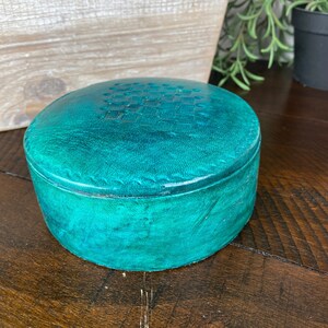 May include: Teal leather trinket box with a textured top. The box is round and has a smooth, polished finish.