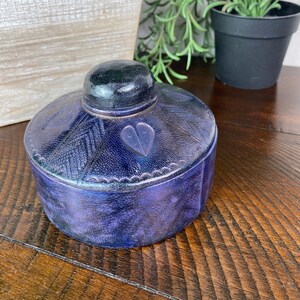 May include: A small, round, blue and purple wooden box with a heart carved into the lid. The box has a decorative edge and a dark wood top.