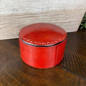 May include: A small, round, red leather trinket box with a black trim and decorative stitching around the top edge.