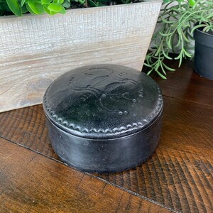 May include: A black leather trinket box with an embossed design. The box is round and has a lid.