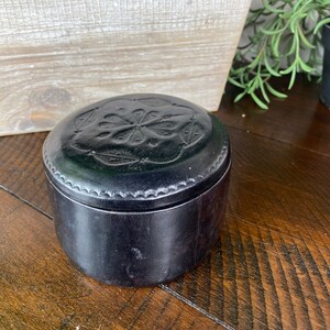 May include: A black leather box with a decorative floral design on the lid. The box is closed and sitting on a wooden surface.