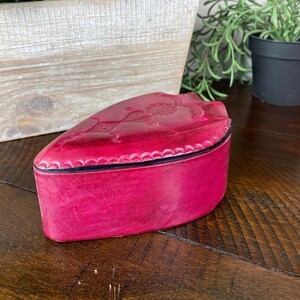 May include: A pink leather heart-shaped trinket box with an embossed design. The box is sitting on a wooden surface.