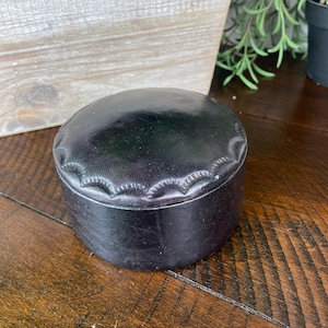 May include: A small, round, black leather box with a decorative stitched design around the top edge. The box is sitting on a wooden surface.