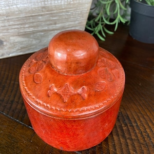 May include: A round, orange leather trinket box with a carved design. The box has a lid with a rounded top.