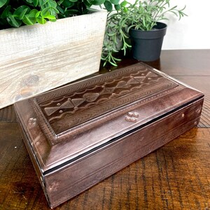 May include: A rectangular, copper-colored box with a detailed, embossed lid featuring geometric patterns. The box is set on a wooden surface, with greenery in the background, suggesting a decorative or storage item.