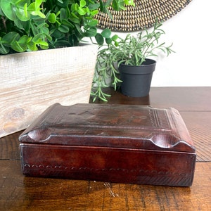 May include: A rectangular, dark brown leather box with a hinged lid. The box features embossed decorative patterns and a scalloped edge. The box is sitting on a wooden surface.