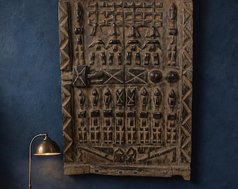 May include: A large, rectangular, weathered wooden door panel featuring intricate, hand-carved geometric patterns and raised relief motifs. The dark brown wood displays a rough, aged texture with a central latch mechanism and symmetrical carvings.