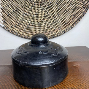 May include: A black leather trinket box with a round shape and a decorative lid. The lid has a raised knob and intricate floral designs.