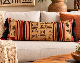 May include: A rectangular lumbar pillow featuring a repeating black and gold geometric Greek key pattern on a red background. The pillow is accented with black and red tassels on both ends, creating a textured and symmetrical appearance.