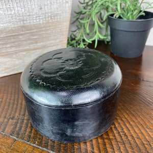 May include: A black leather round box with a decorative design on the top. The box is sitting on a wooden surface.