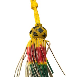 May include: A decorative tassel with a yellow braided top and a round, black and yellow patterned center. The tassel features yellow, red, and green leather strips, with natural-colored leather strips at the bottom.