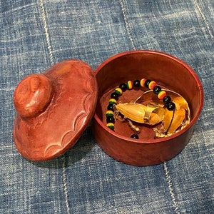 May include: A small, round, red leather box with a lid. The lid has a carved design and a knob on top. The box is open and contains a gold necklace, a gold ring, and a black and yellow beaded bracelet.