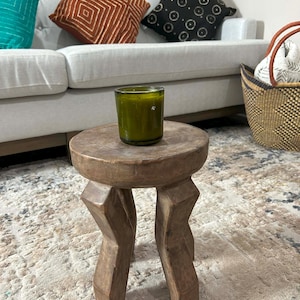 Puede incluir: Un pequeño taburete redondo de madera con tres patas talladas. Un portavelas de vidrio verde se encuentra encima. El fondo incluye un sofá blanco con cojines decorativos en verde azulado, óxido y negro, y una cesta tejida.
