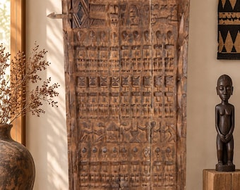 May include: A tall, rectangular, weathered brown wooden door panel mounted on a black metal stand. The surface is intricately carved with rows of stylized human figures, geometric patterns, and small hut motifs, featuring a prominent latch mechanism.