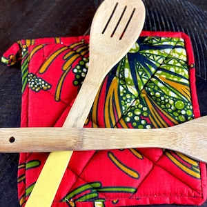 May include: Two wooden kitchen utensils, a slotted spoon and a spatula, are placed on a red quilted hot pad with a colorful African print design.