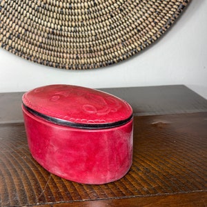 May include: A red leather oval-shaped trinket box with a black trim and a decorative design on the lid.