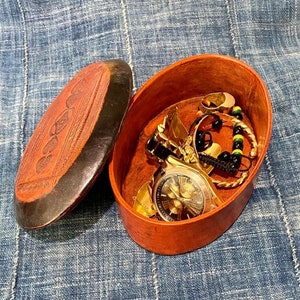 May include: A brown leather oval box with a lid, open to reveal a gold watch, a gold bracelet, and a black beaded bracelet with green and yellow beads.