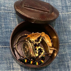 May include: A round wooden box with a lid, open to reveal a gold watch, gold bracelets, and a black beaded bracelet with green and yellow accents.