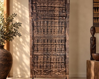 May include: A tall, rectangular, dark brown wooden panel featuring intricate hand-carved relief patterns of human figures and geometric shapes. The panel is mounted vertically on a simple, flat, black metal base, showcasing a weathered texture.