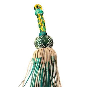 May include: A decorative tassel featuring a woven green and white ball, a braided yellow and green top, and a fringe of tan leather strips with green tips. The tassel is designed with a loop at the top.
