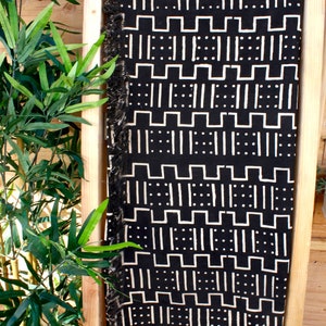 K&ouml;nnte beinhalten: Schwarz-wei&szlig;e Tagesdecke mit geometrischem Muster. Die Decke hat einen Fransenrand und wird auf einem Holzregal pr&auml;sentiert.