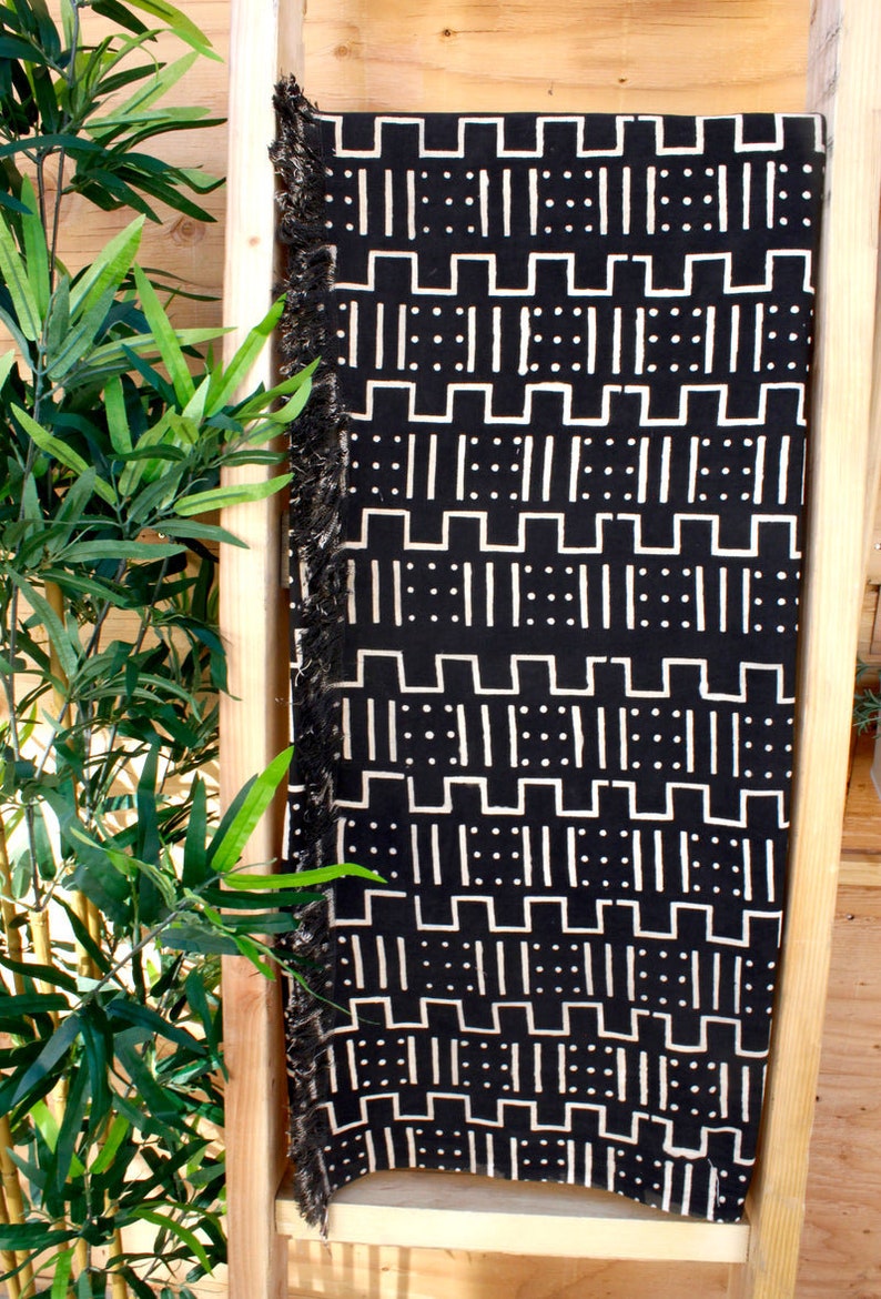 K&ouml;nnte beinhalten: Schwarz-wei&szlig;e Tagesdecke mit geometrischem Muster. Die Decke hat einen Fransenrand und wird auf einem Holzregal pr&auml;sentiert.