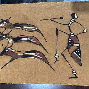 Może przedstawiać: Brązowy wiszący obraz wykonany z tkaniny z czarno-białą stylizowaną ilustracją osoby tańczącej z trzema zwierzętami. Osoba ma na sobie wzorzystą spódnicę i naszyjnik. Zwierzęta są stylizowane i mają długie szyje i ogony.