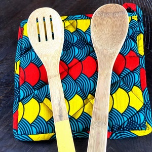 May include: Two wooden kitchen utensils, a slotted spoon and a solid spoon, are placed on a colorful fabric hot pad with a blue, yellow, and red pattern.