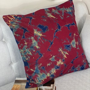 May include: A square decorative pillow with a tie-dye pattern. The pillow features a deep red background with blue and yellow accents. The pillow is resting on a light gray couch.