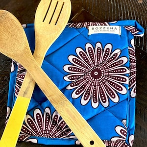 May include: Two wooden spoons with yellow and brown handles are placed on top of a blue quilted hot pad with a brown and white floral pattern. The hot pad has a white label with the text "BOZZENA www.bozzena.com".