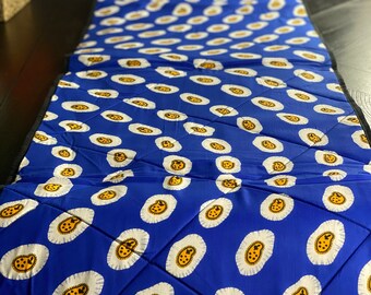 Peut inclure: Un chemin de table bleu avec un motif d'œufs stylisés blancs et jaunes. Le chemin de table a une bordure noire et est matelassé.