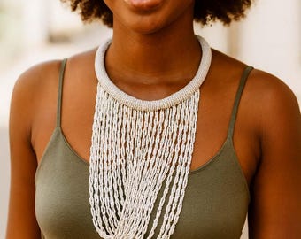 May include: A white beaded statement necklace with a wide collar and cascading fringe. The necklace is made of small, round beads and hangs down the chest. The model is wearing a green tank top.