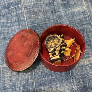 May include: A red leather box with a floral design on the lid. The box is open and contains a gold watch with a black face, a gold bracelet, and a gold necklace.