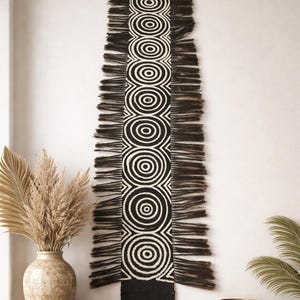 May include: A long, rectangular wall hanging with a black and white geometric design. The central pattern features concentric circles, framed by a black border and long, dark fringe. The artwork is displayed against a white wall, suggesting a modern home decor theme.