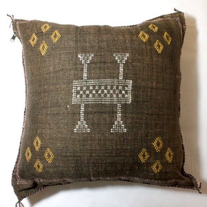 Puede incluir: Una almohada de tiro marrón con un patrón geométrico blanco y dorado. La almohada tiene una cremallera.