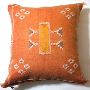 May include: A square orange throw pillow with a geometric pattern in white, yellow, and blue thread. The pillow has a zipper closure.