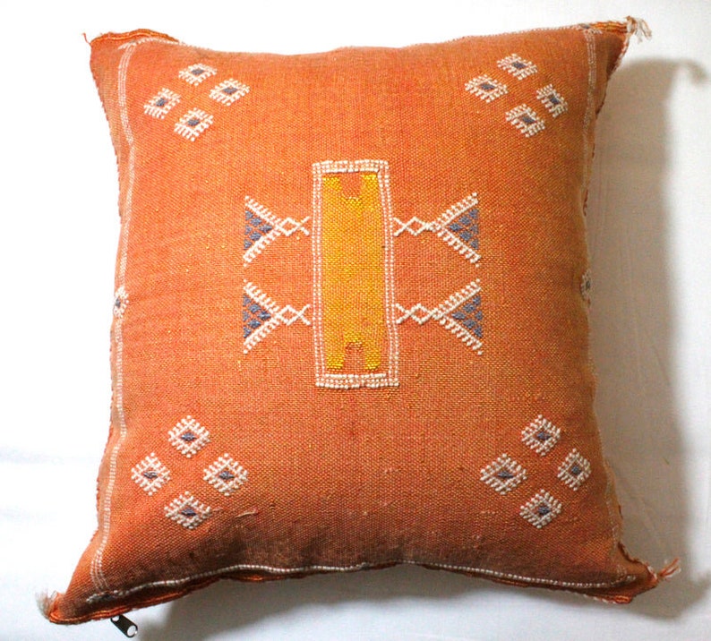 Op de afbeelding: Een vierkant oranje kussen met een geometrisch patroon in wit, geel en blauw garen. Het kussen heeft een rits.