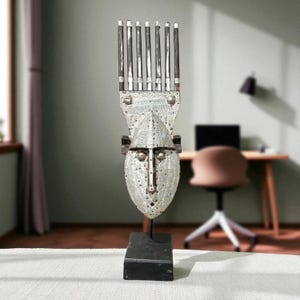 Pode incluir: Escultura de máscara tribal prateada com base retangular. A máscara apresenta um rosto detalhado e uma estrutura semelhante a um pente no topo. A escultura é exibida em uma superfície branca.