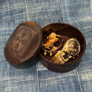 May include: A brown leather jewelry box with an embossed floral design on the lid. Inside, a gold-toned watch, bracelets, and rings are visible. The box is open, revealing the contents.