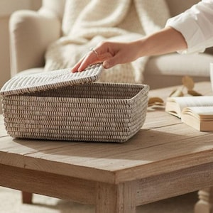 Puede incluir: Una cesta de almacenamiento rectangular tejida con tapa, en color neutro, está sobre una mesa de madera. La tapa está parcialmente abierta, revelando el interior. La cesta está junto a un libro abierto y una taza blanca.